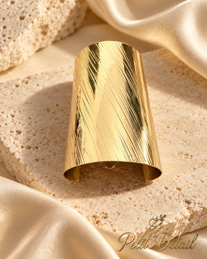 Wide gold-tone hair cuff with diagonal brushed texture styled on beige stone and satin