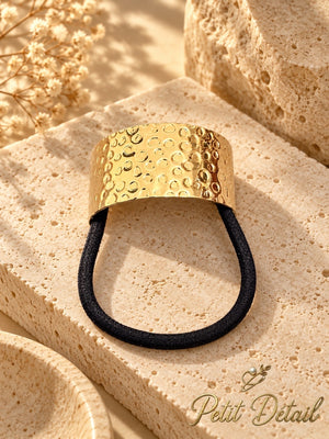 Embossed circular bubble-pattern gold-tone hair cuff with black elastic, photographed on beige travertine stone