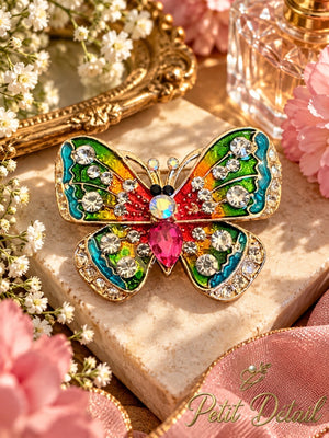 Rainbow enamel butterfly brooch with clear crystal-look accents and vibrant pink center styled on beige stone with flowers and pink ribbon