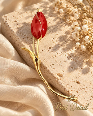 Red enamel long-stem tulip brooch displayed on beige stone beside ivory fabric and tiny white flowers