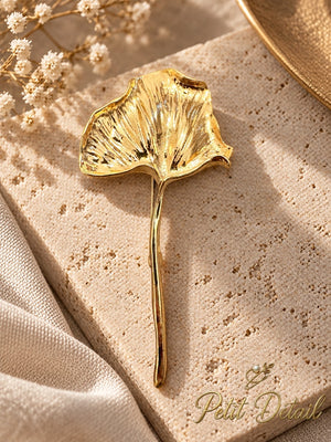 Textured gold-tone long-stem ginkgo leaf brooch displayed on beige stone with cream textile and delicate white flowers