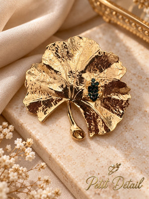 Textured gold-tone leaf brooch with deep green crystal-look details displayed on beige stone beside ivory satin and white flowers