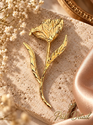Sculptural gold-tone open flower brooch with two slender leaves displayed on travertine beside ivory satin and white flowers